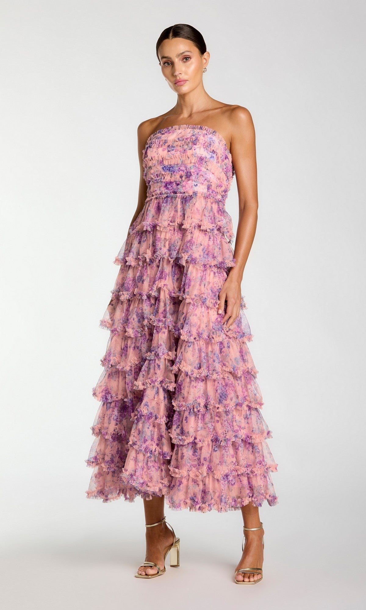A woman wearing the Mac Duggal 8420 Tea-Length Party Dress in Orchid Multi—a strapless, ankle-length ruffle tiered dress—paired with gold high-heeled sandals, stands against a plain light background.